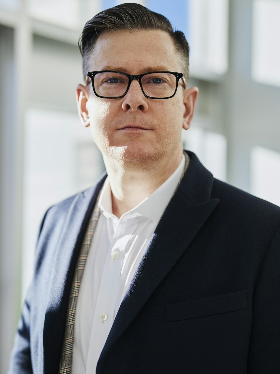 A man in glasses and a navy blazer looking toward the camera.